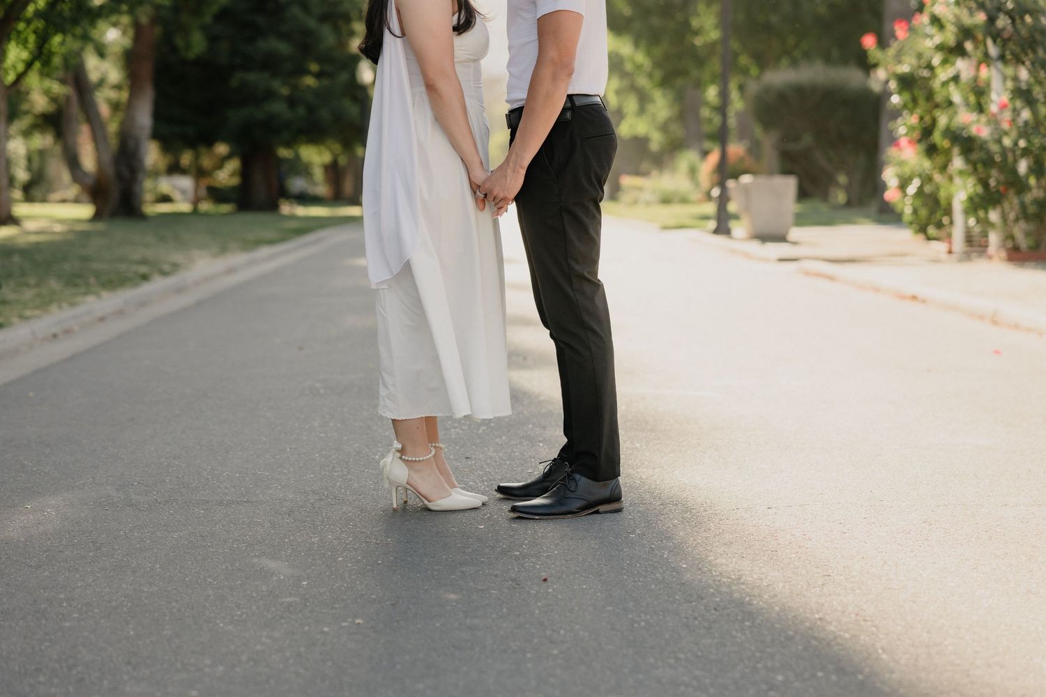 Couple holding hands in wedding attire on a sunlit park path, bride in white dress and groom in black pants.