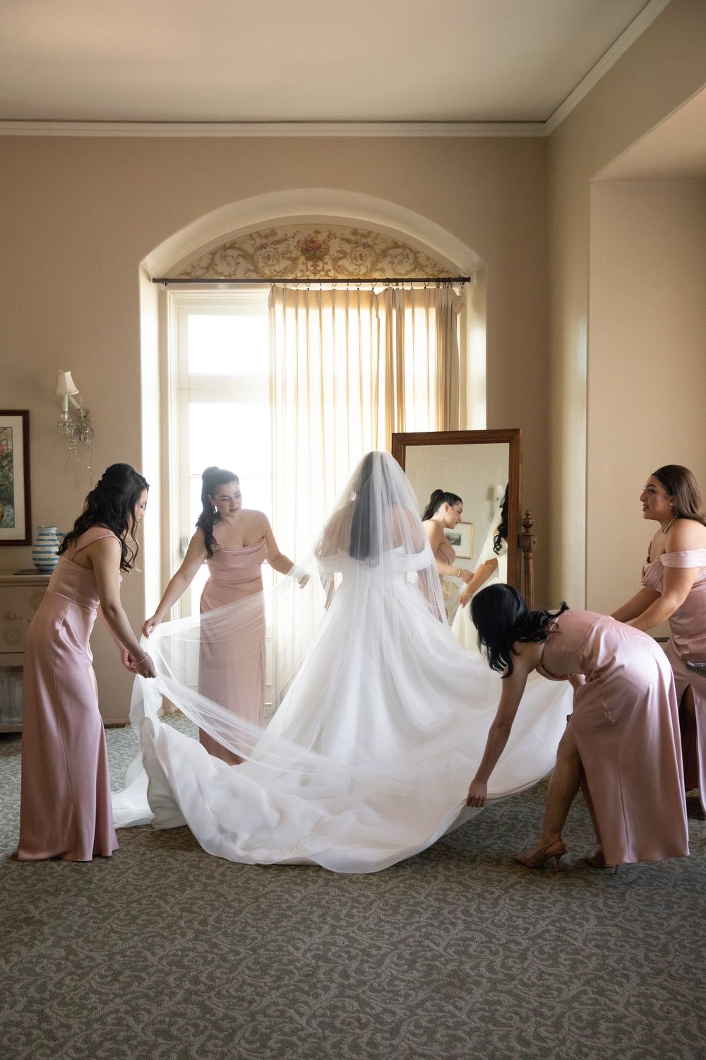 Bridesmaids in pink gowns help bride arrange her white wedding dress and veil in an elegant hotel room.