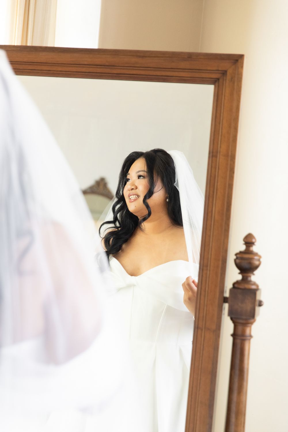Bride in white wedding dress and veil admiring her reflection in a wooden floor mirror while getting ready.
