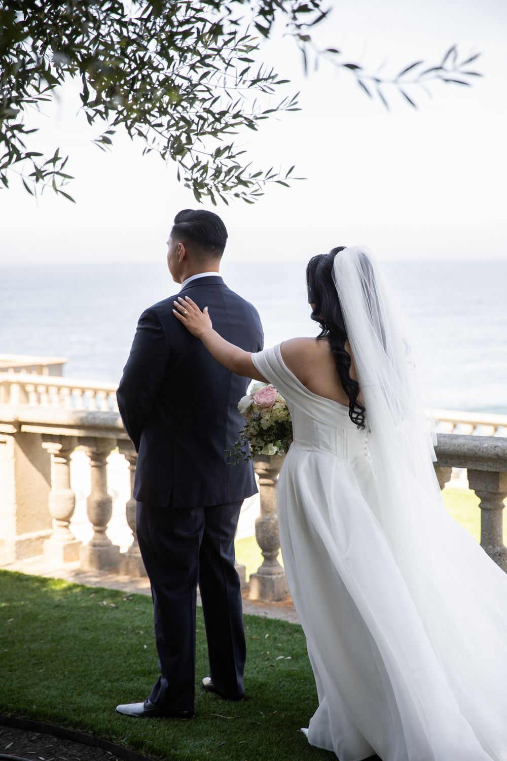 Bride and groom stand together overlooking the ocean on a balcony with stone balustrade at their wedding.