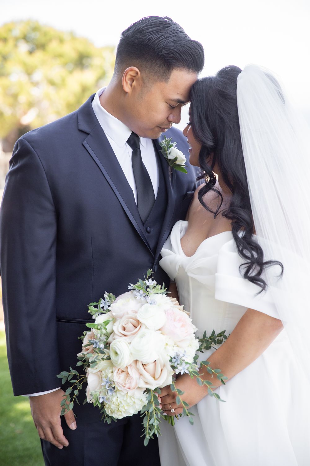 Bride and groom touching foreheads tenderly outdoors, bride holding white and blush bouquet, groom in navy suit.