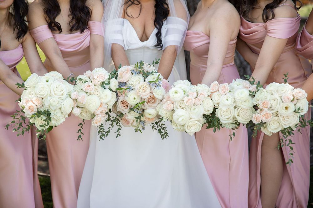 Bride and bridesmaids in pink off-shoulder dresses holding lush white and blush rose bouquets at a wedding ceremony.