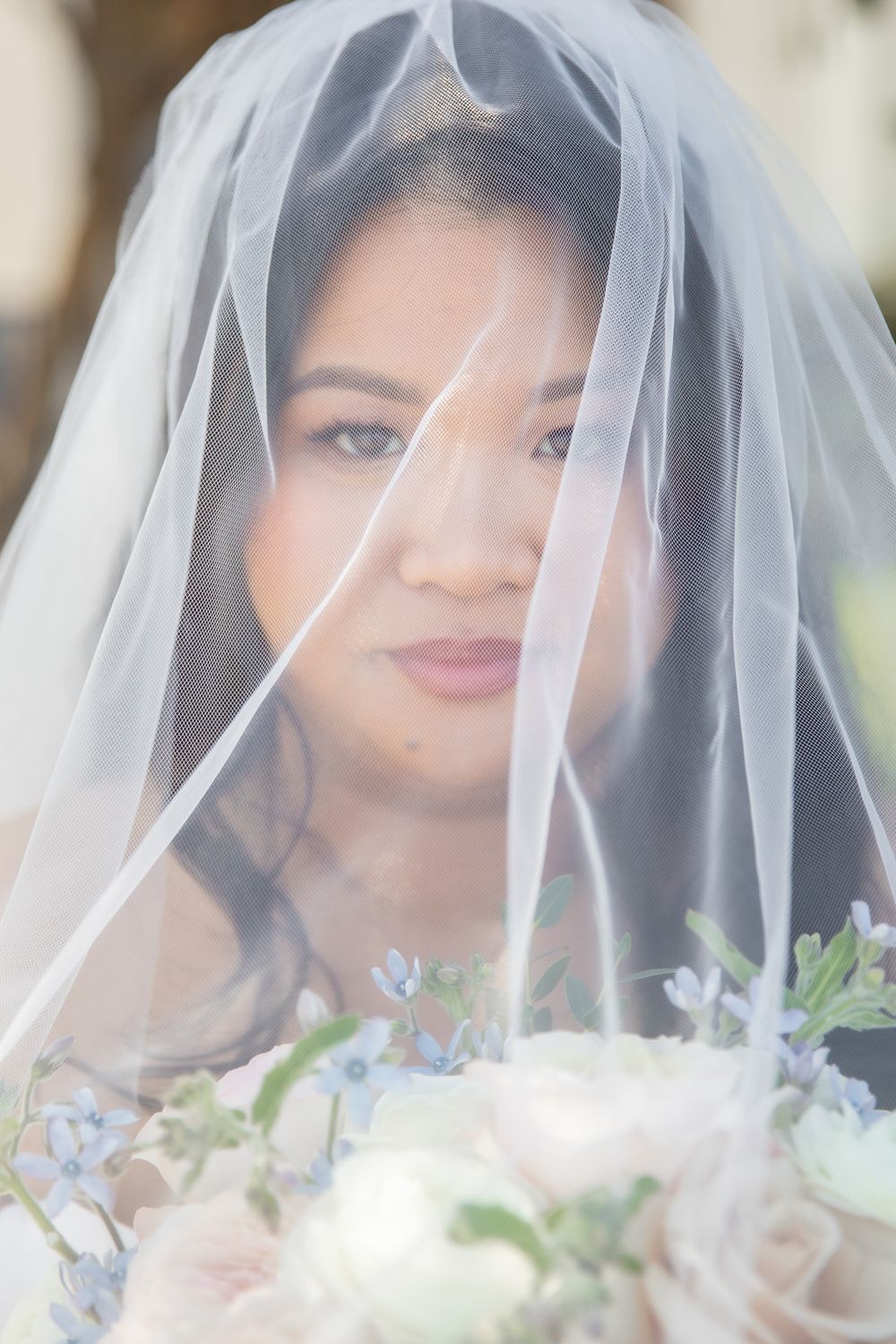 Close-up portrait of Asian bride wearing a sheer veil holding a bouquet of white and blue flowers.