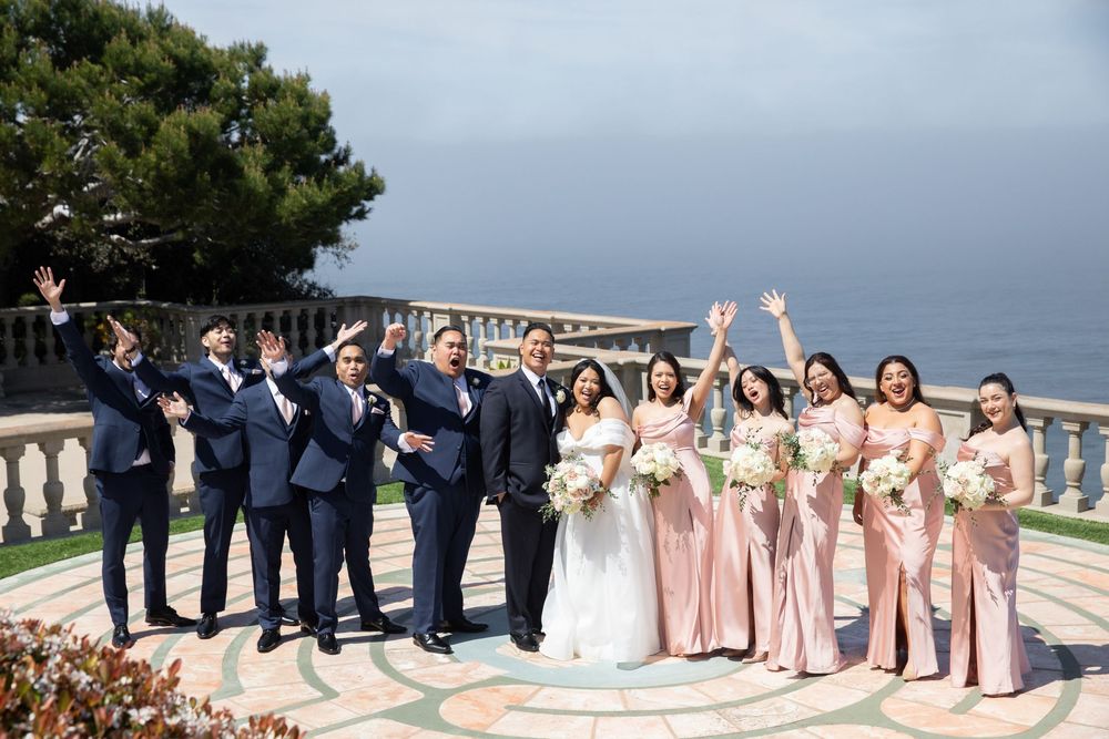 Wedding party celebrates on terrace with ocean view, groomsmen in navy suits and bridesmaids in blush pink gowns.