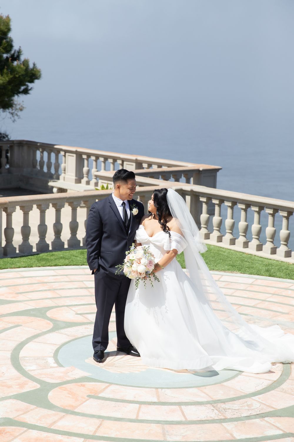 Bride and groom share a romantic moment on a terrace overlooking the ocean, bride holding a white floral bouquet.