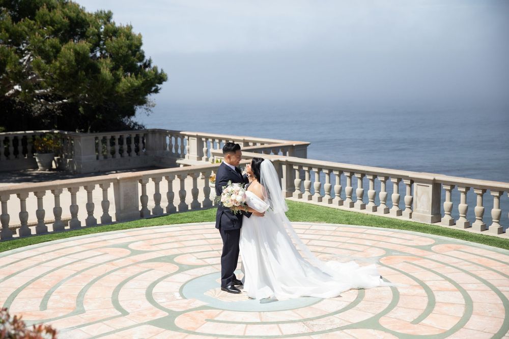 Bride and groom sharing romantic moment on scenic ocean-view terrace, bride in white gown with long cathedral veil.