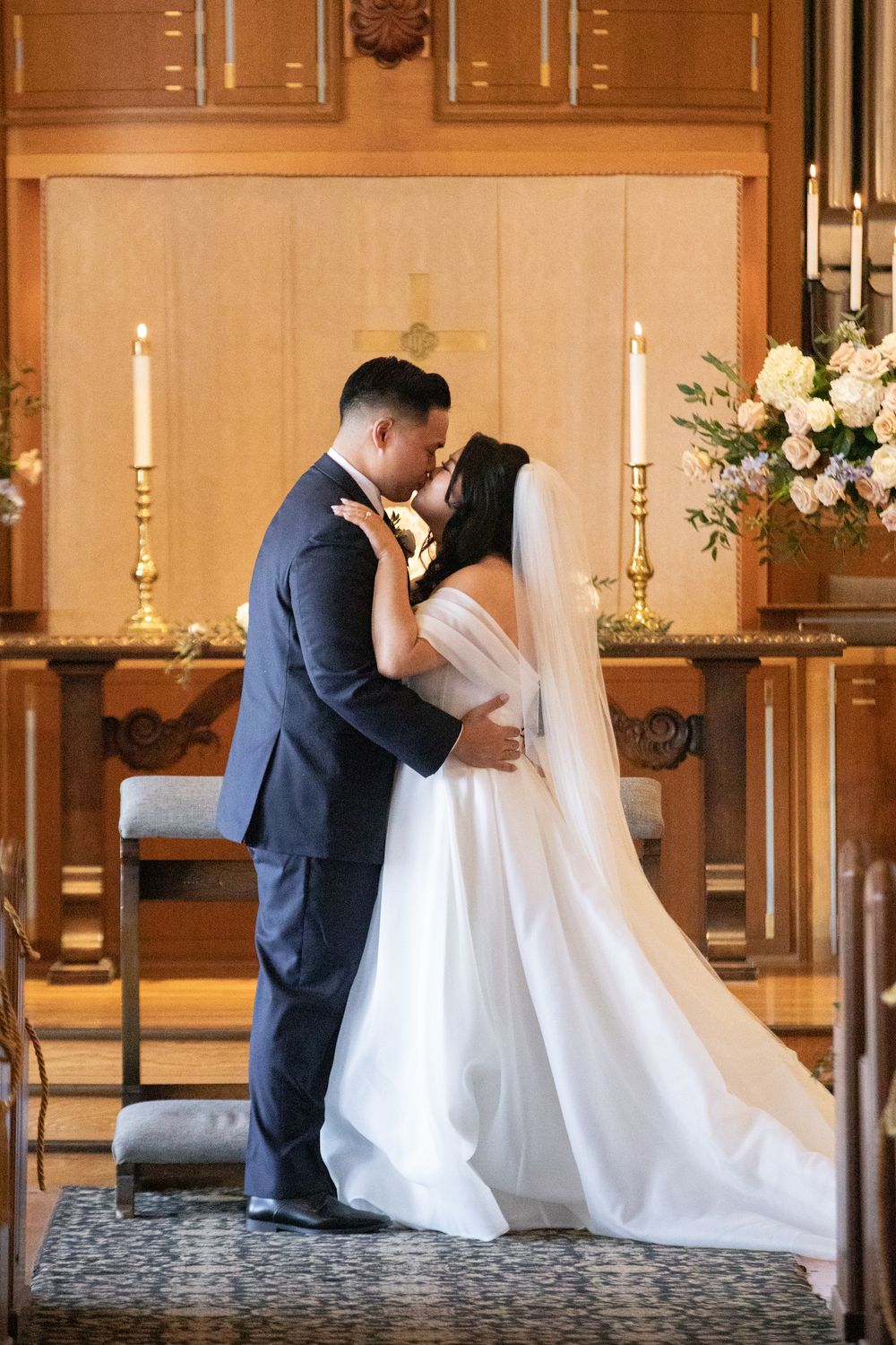 Bride and groom share first kiss at church altar with candles and floral arrangements during wedding ceremony.