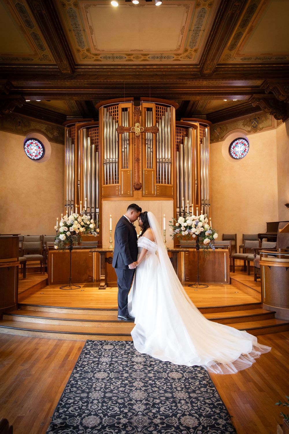 Bride and groom embrace before grand pipe organ altar with floral arrangements in elegant church wedding.