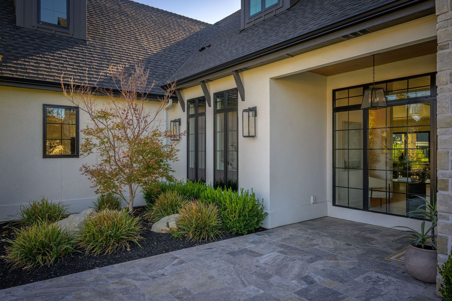 Modern home exterior with stone facade, black-framed windows, wall lanterns, and manicured landscaping on a patio.