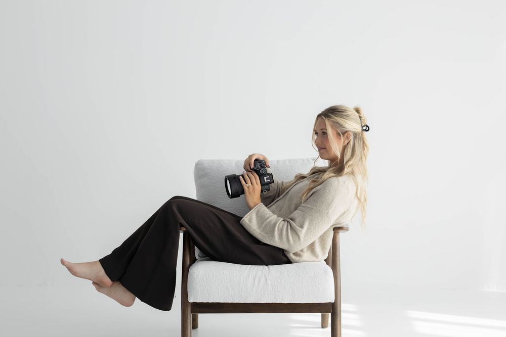 Blonde woman sitting in a white chair reviewing photos on a camera in a bright minimalist studio setting.