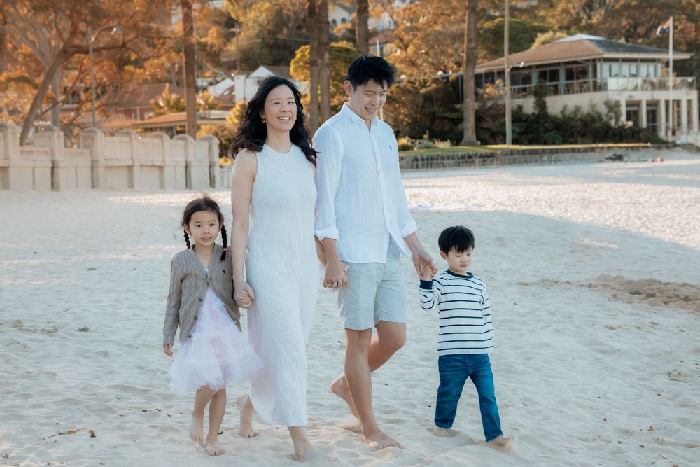 Family of four walking on sandy beach, parents holding children's hands, wearing white outfits, trees and building in background.