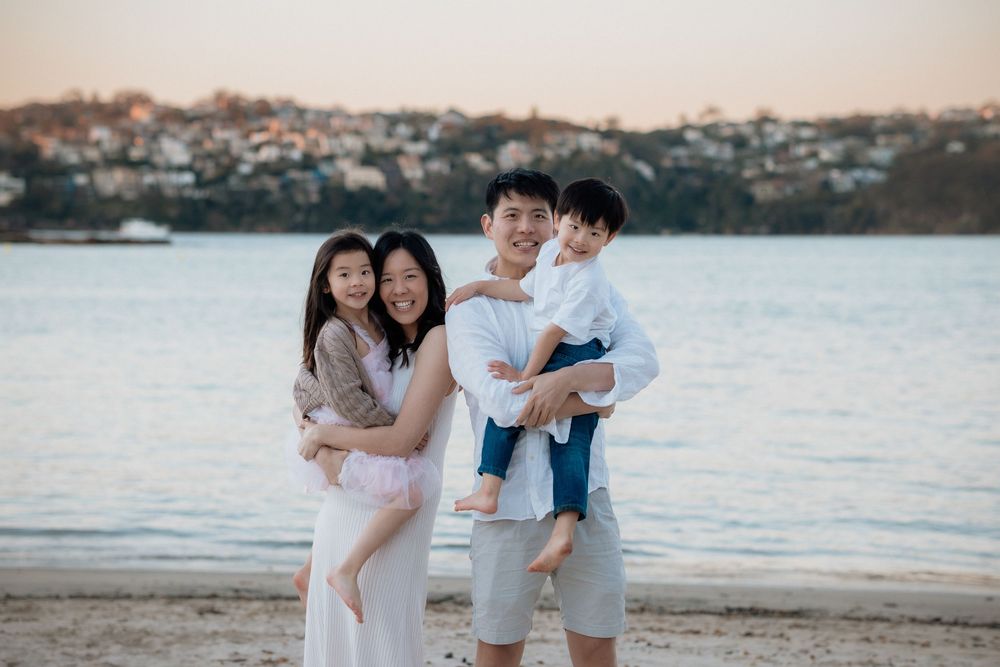 Happy Asian family of four posing on a sandy beach at sunset with a scenic waterfront town in the background.