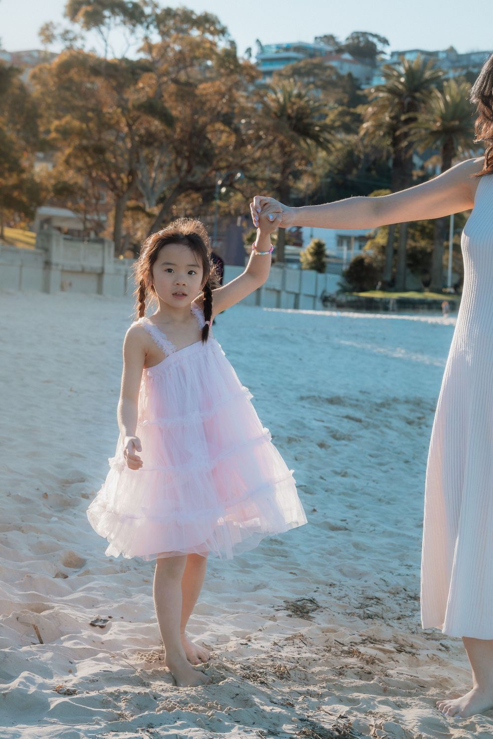 A young girl in a pink ruffled dress holds hands with a woman on a sandy beach surrounded by palm trees.