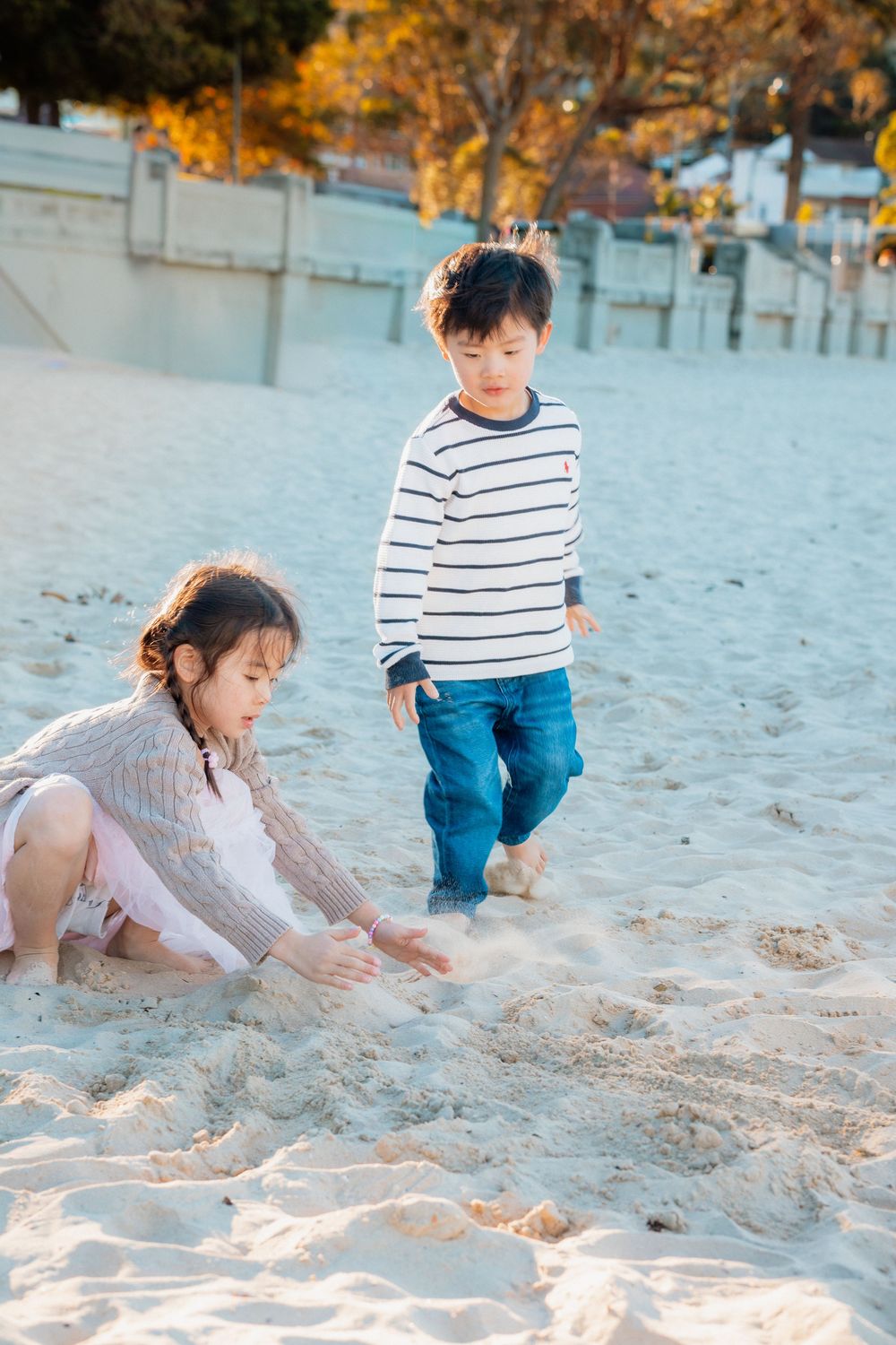 Two Asian children playing in white sand at a beach, girl sitting while boy stands nearby wearing striped shirt and jeans.