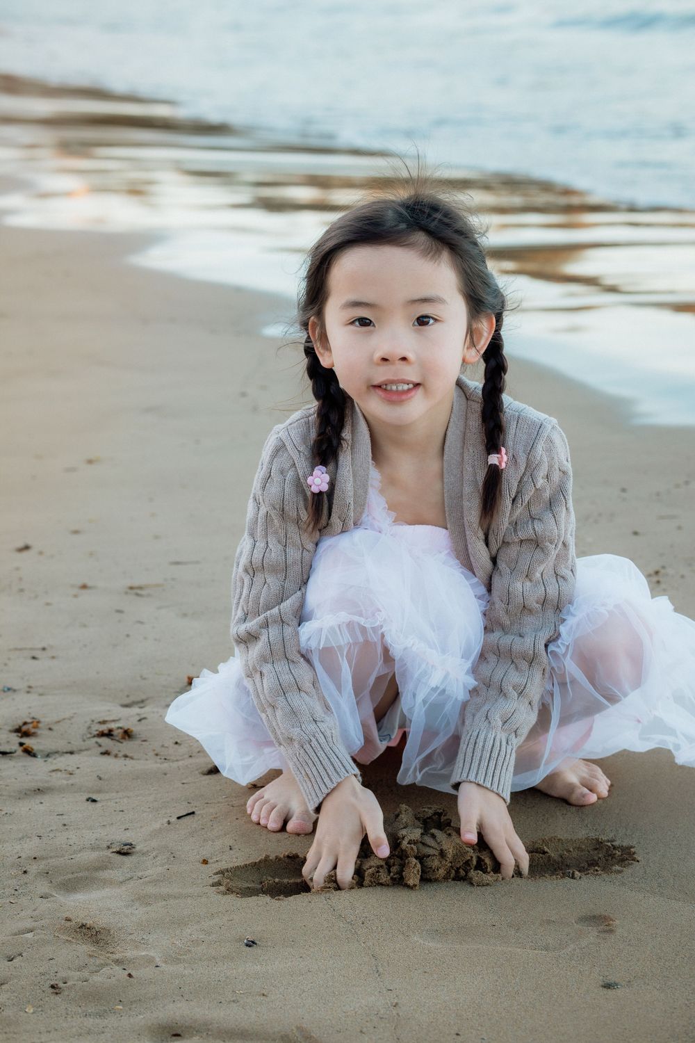 Young Asian girl with pigtails playing in sand on beach, wearing knit cardigan and white tulle skirt.