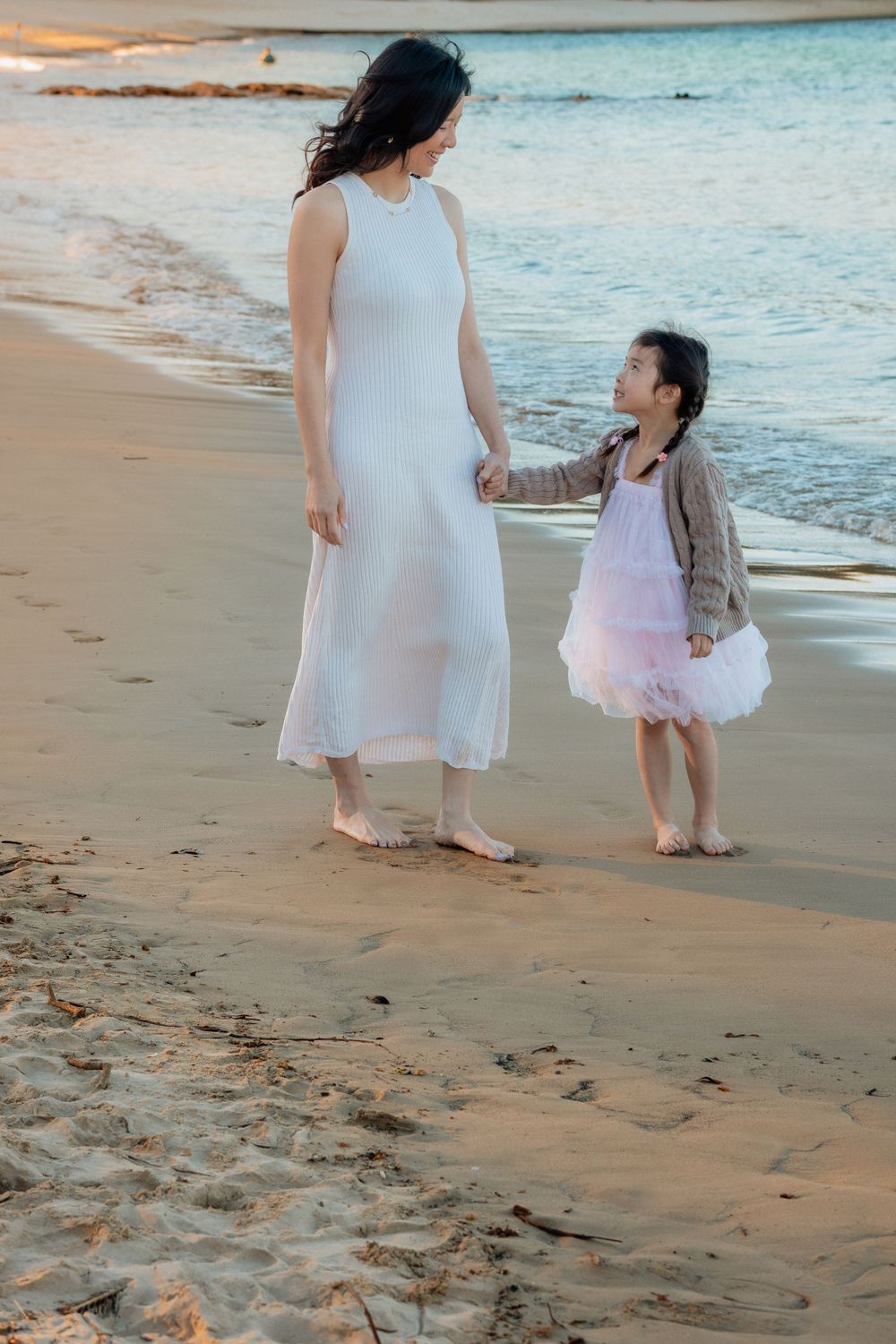 A mother in a white dress holds hands with her young daughter in a pink dress while walking barefoot on a sandy beach.