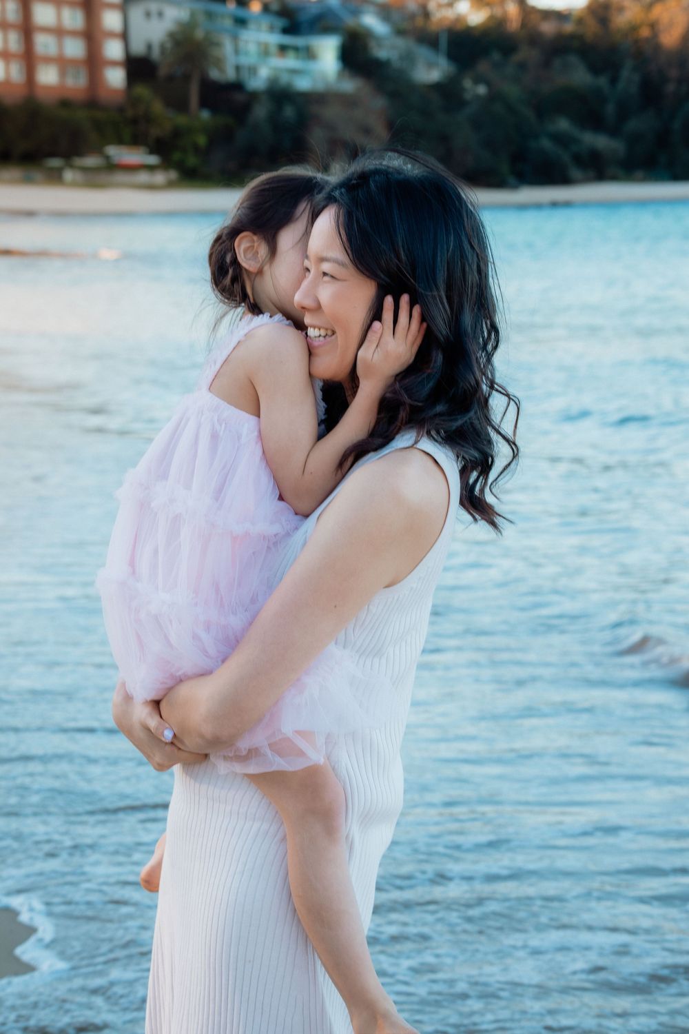 A mother laughs joyfully while holding her young daughter on a beach at sunset, both wearing white and pink dresses.