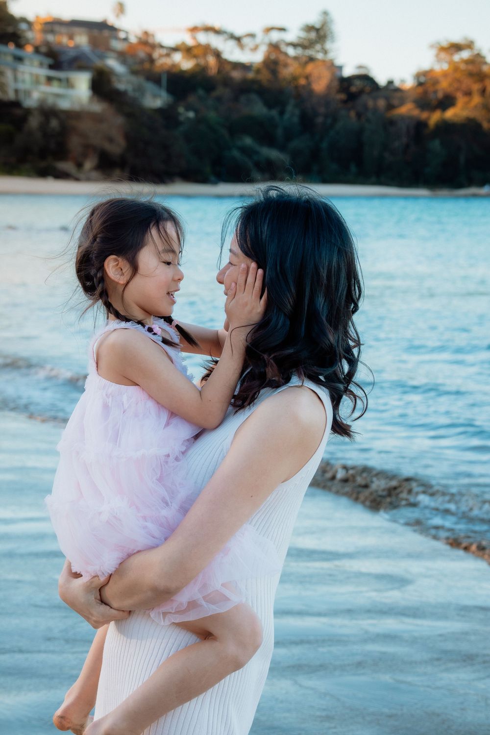 A mother holds her young daughter on a beach at sunset, sharing a tender moment near calm blue water.