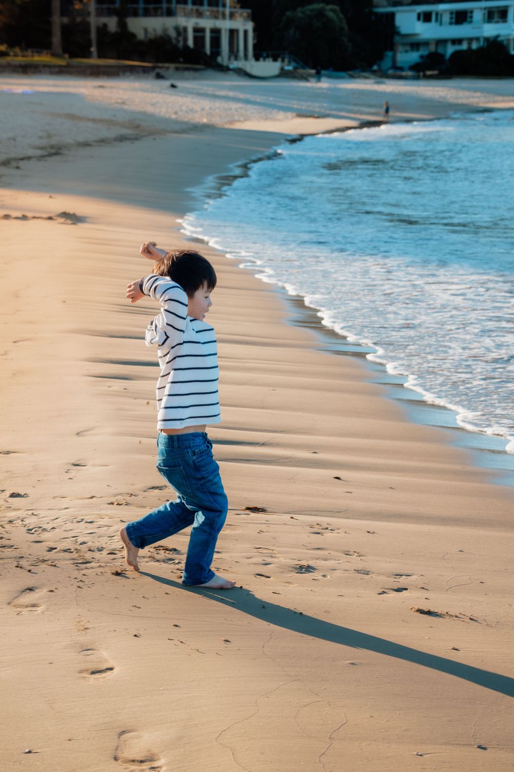A young boy in a striped shirt and jeans walks barefoot along a sandy beach at sunset.