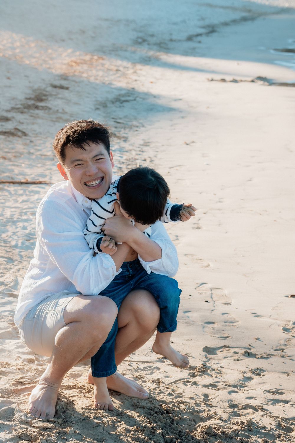 Happy Asian father laughing while hugging young son on sandy beach in white shirt and shorts.