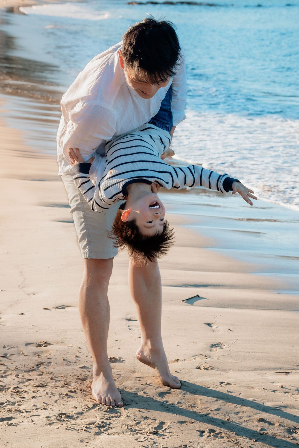 A father playfully holds his laughing child upside down on a sunny sandy beach with blue ocean waves in the background.