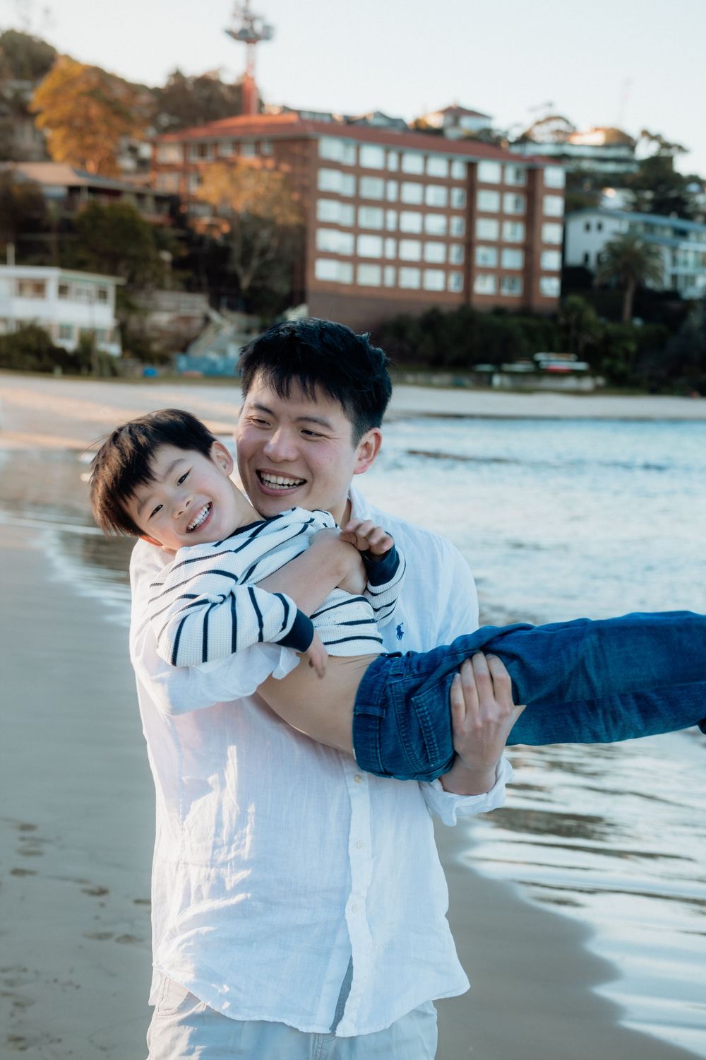 A smiling man carries a laughing child on a sandy beach with buildings and trees in the background.