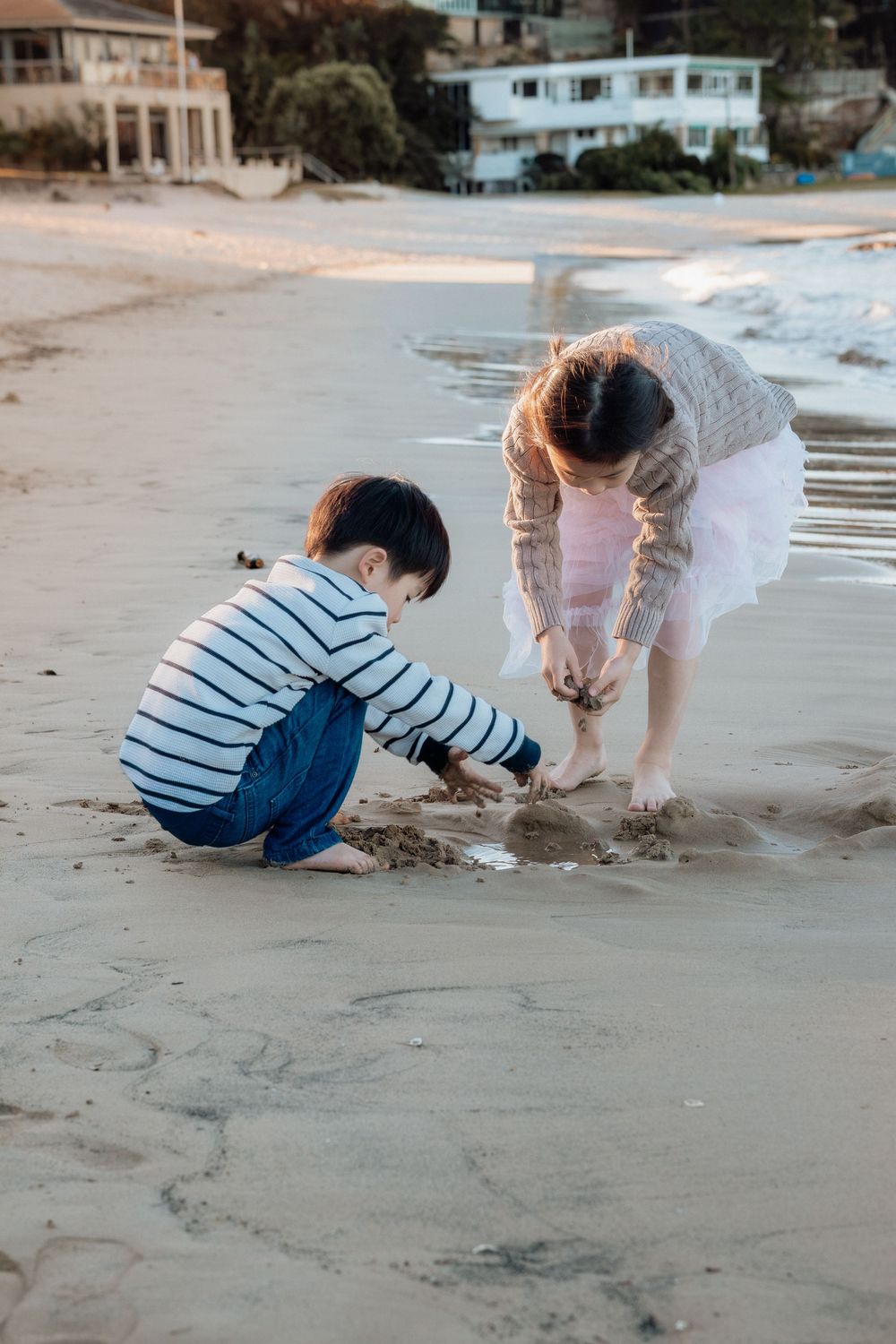 Two young children playing in the sand on a beach at sunset, with houses and trees visible in the background.