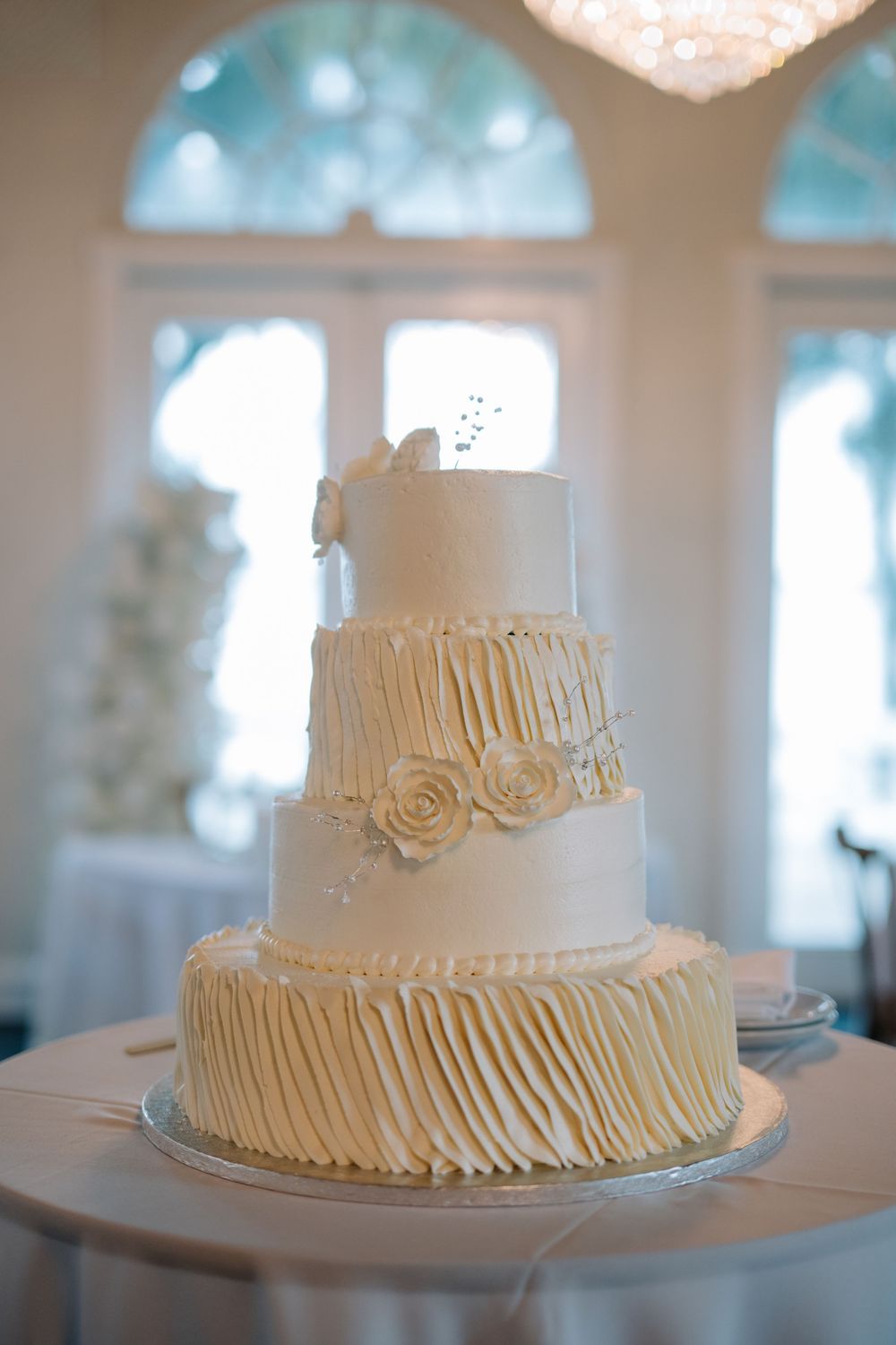 Elegant three-tier white wedding cake with ruffled frosting, rosettes, and butterfly topper displayed in a bright reception hall.