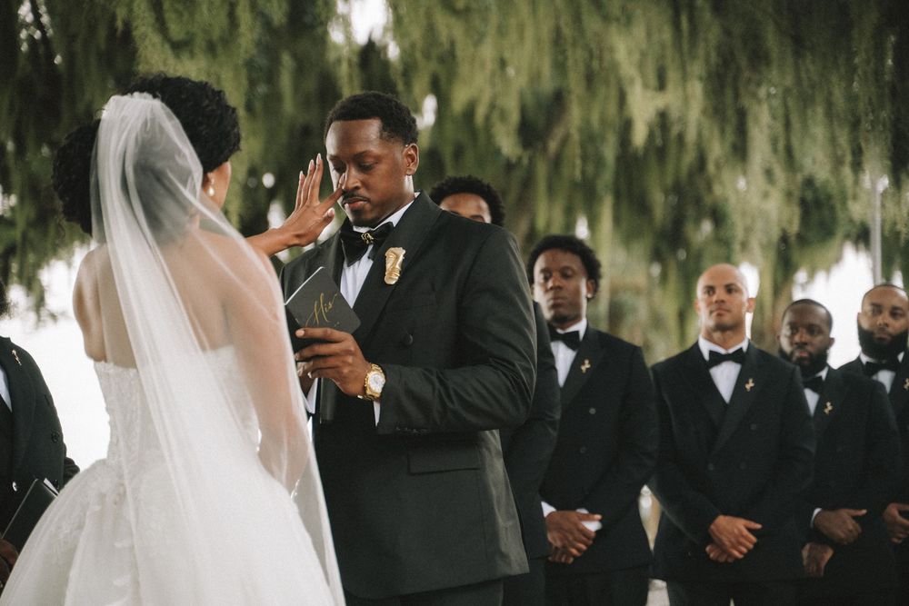 Groom reading vows to bride during outdoor wedding ceremony with groomsmen standing in background.