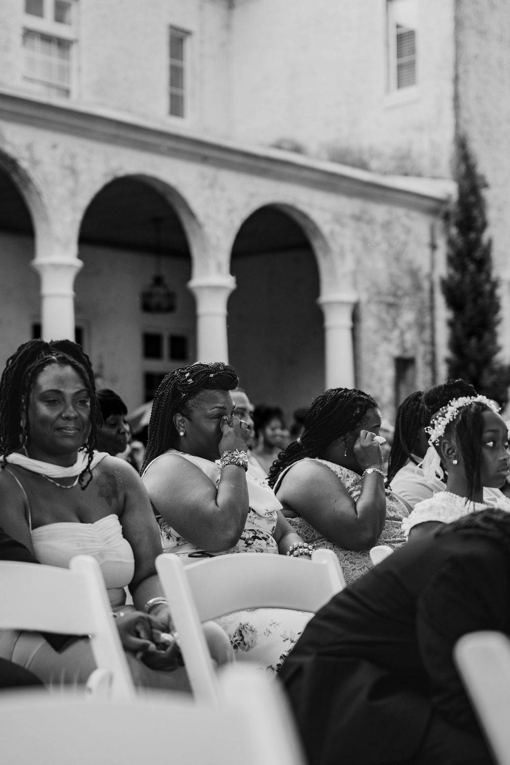 Black and white photo of emotional wedding guests seated outdoors at a ceremony, some wiping tears, wearing formal attire.