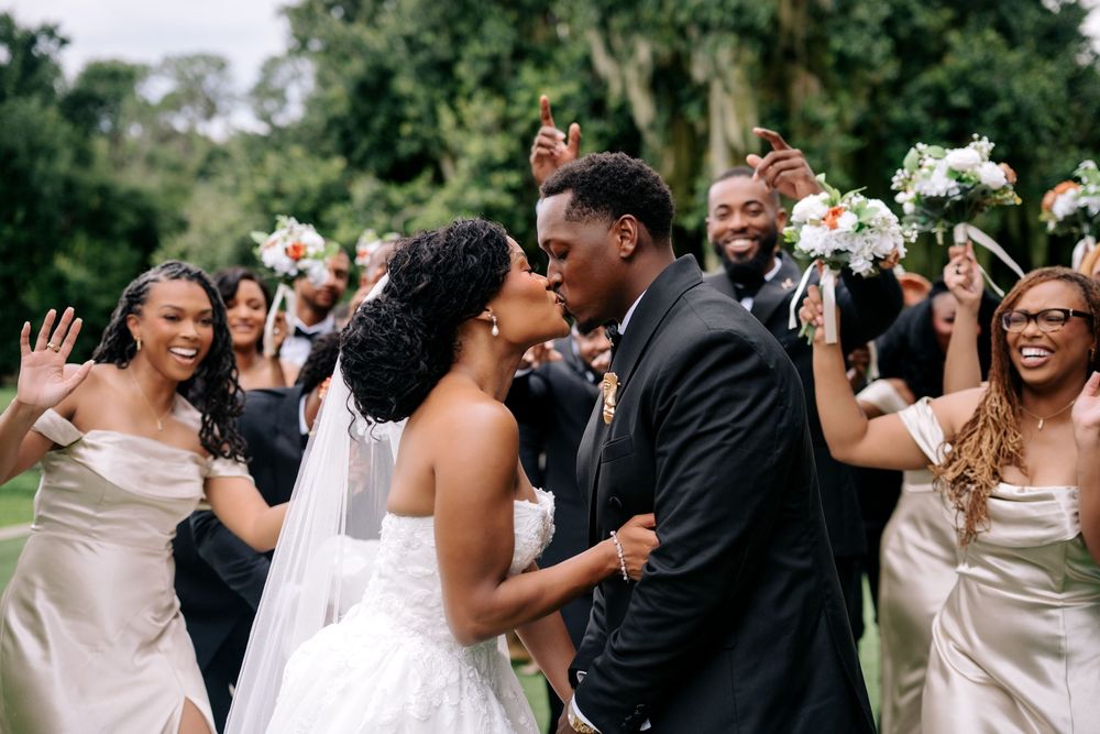 A joyful wedding couple shares a kiss surrounded by cheering bridesmaids and groomsmen holding bouquets outdoors.