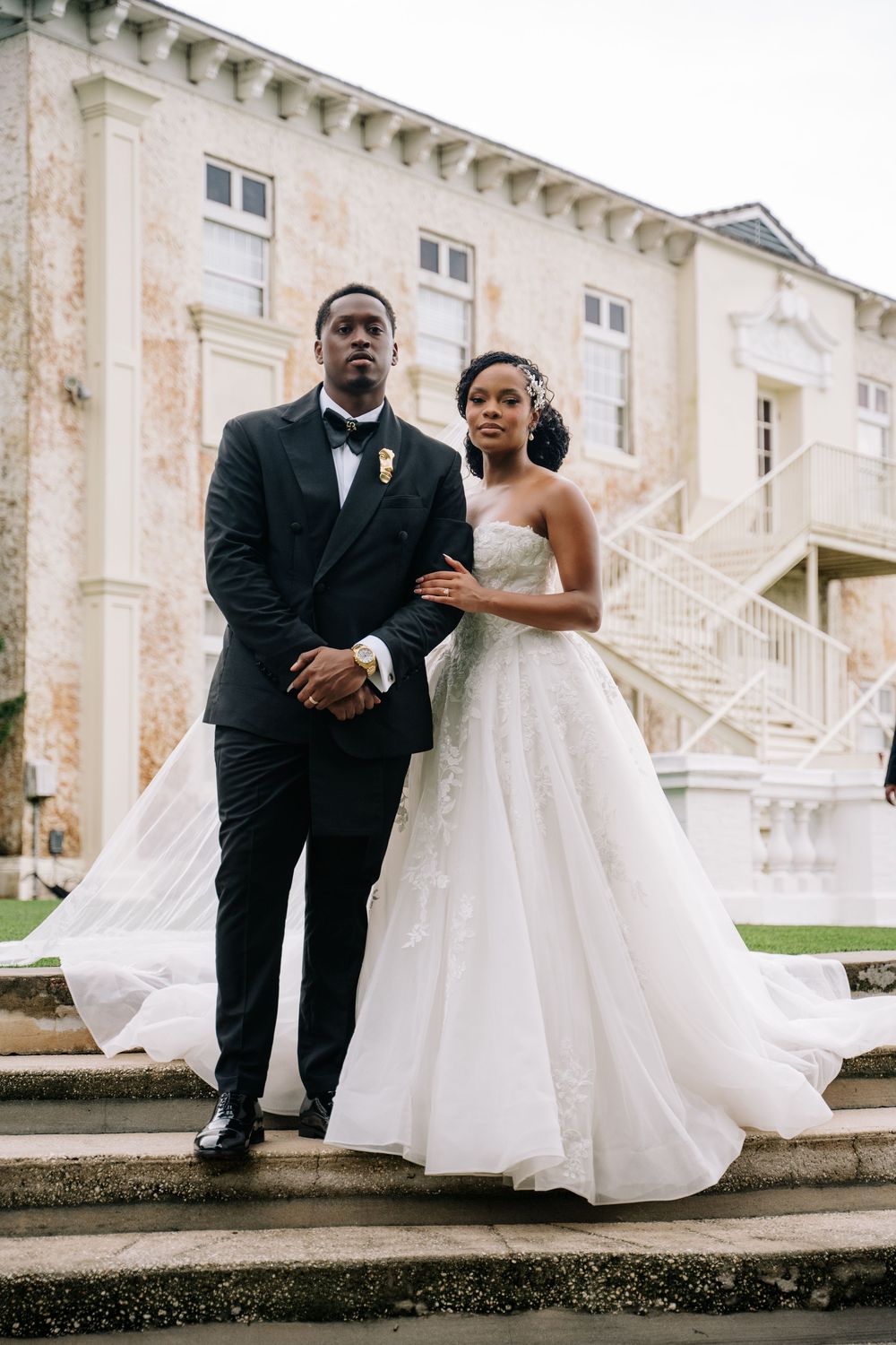 Elegant bride and groom pose outside grand estate, she in strapless ball gown, he in classic black tuxedo.