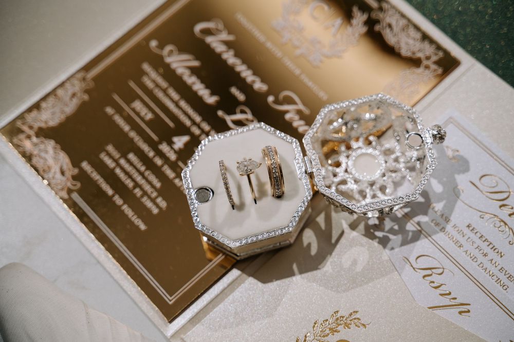 Elegant wedding rings displayed in a white jeweled box atop gold mirror wedding invitations with floral details.