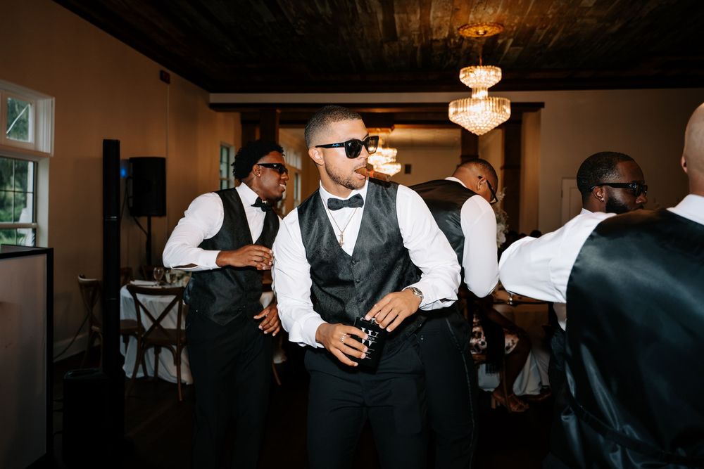 Groomsmen in black vests and bow ties dancing at a wedding reception in an elegant venue.