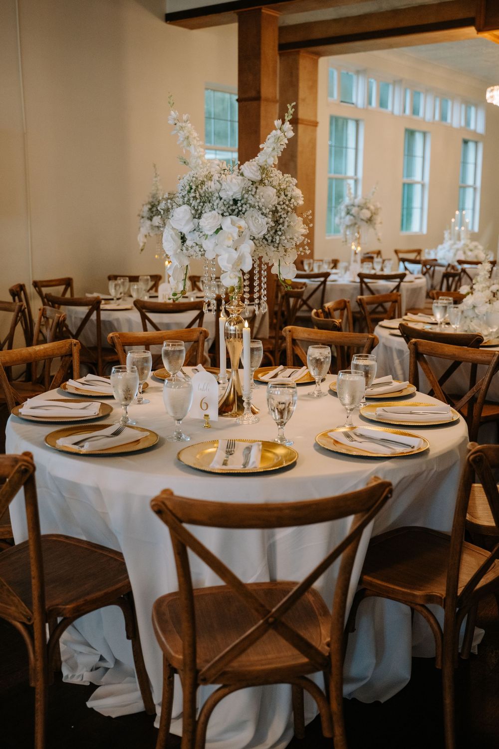 Elegant wedding reception tables with white linens, gold chargers, tall floral centerpieces, and rustic wooden cross-back chairs.