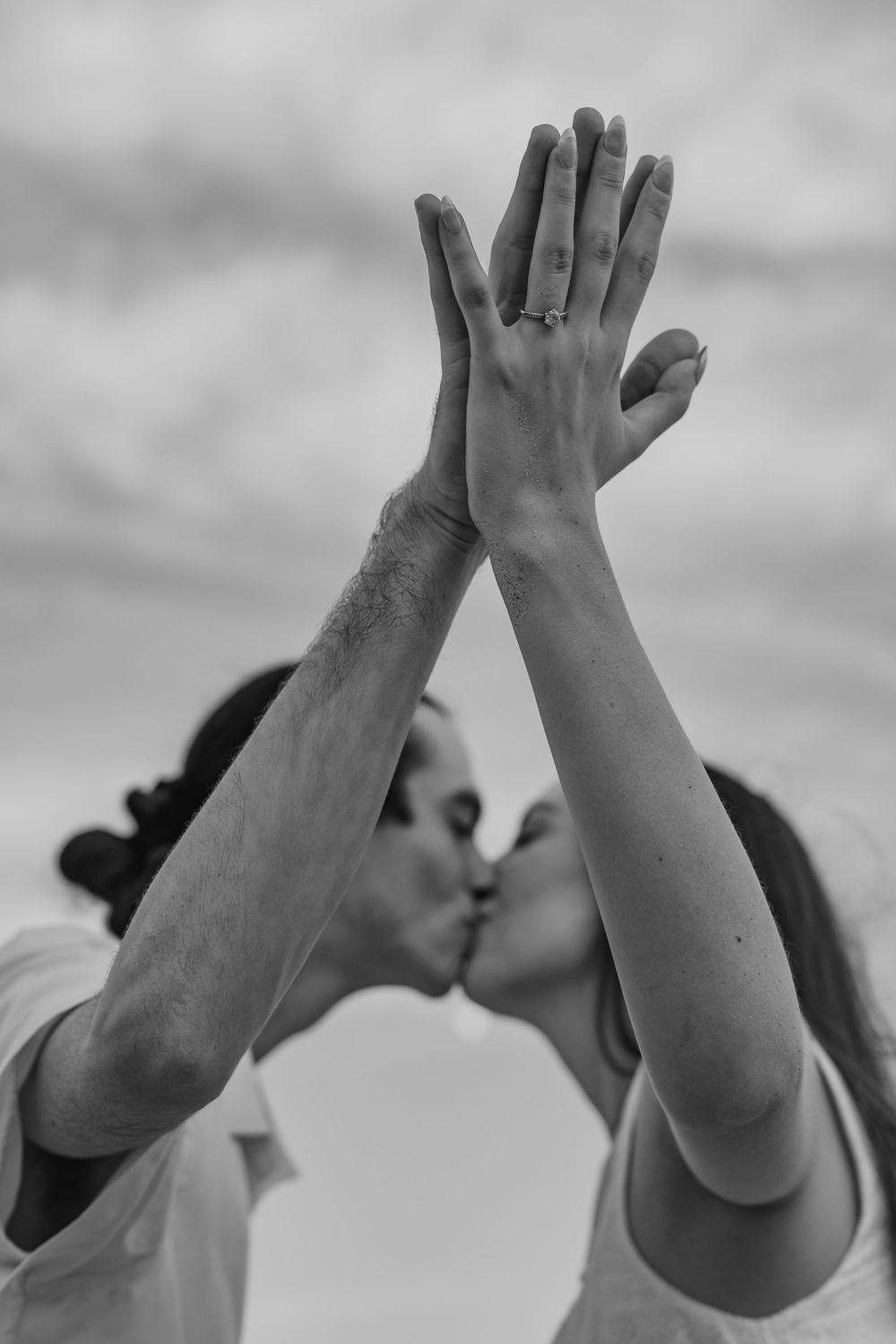 Black and white photo of a couple kissing while raising their hands together, showing an engagement ring.