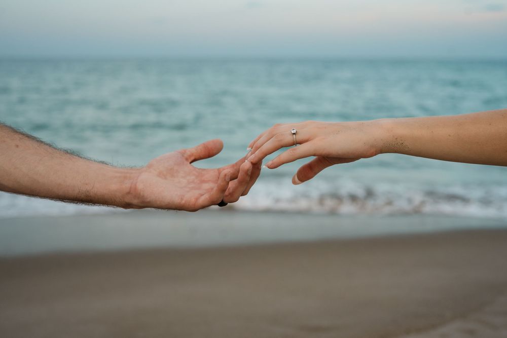 Two hands reaching toward each other on a beach with ocean waves in the background.