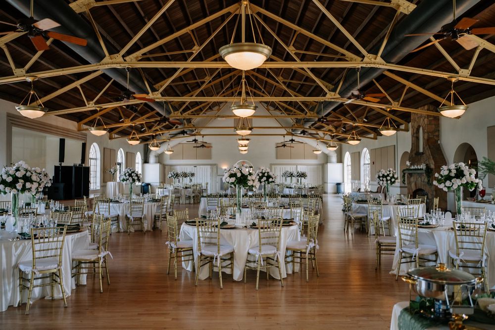 Elegant wedding reception hall with vaulted wood ceiling, round tables with white linens, gold chairs, and floral centerpieces.