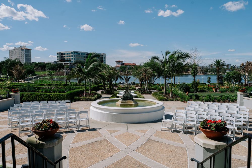 Elegant outdoor wedding ceremony setup with white chairs, fountain, palm trees, and waterfront city views under a blue sky.