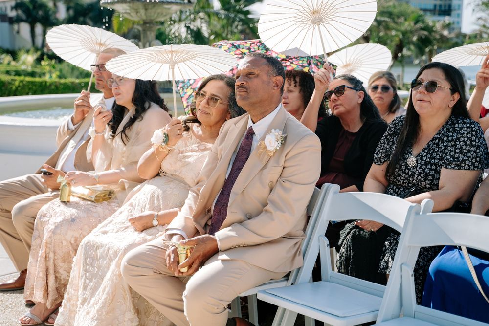 Guests seated at an outdoor wedding ceremony, holding white parasols for shade in a sunny tropical setting.