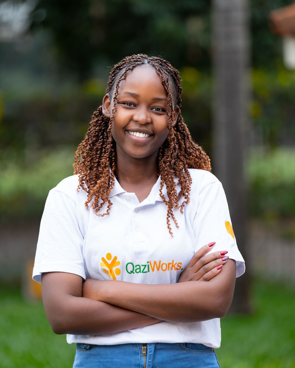 Smiling young woman with braided hair wearing a QaziWorks white polo shirt with arms crossed outdoors.