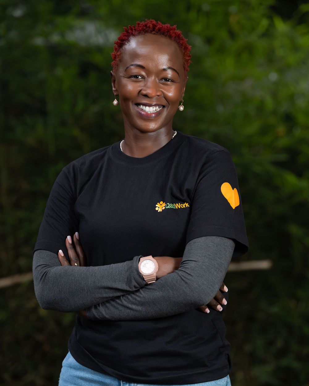 Smiling woman with red hair wearing a QaziWorks black t-shirt standing with arms crossed outdoors.