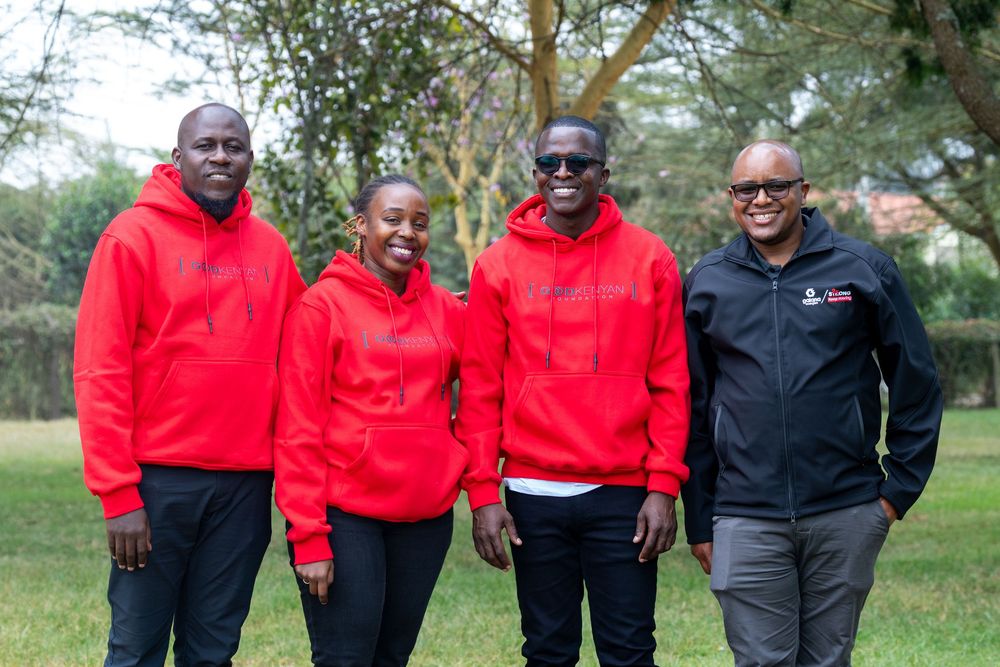 Four people in matching red hoodies pose together outdoors on green lawn with trees in background.