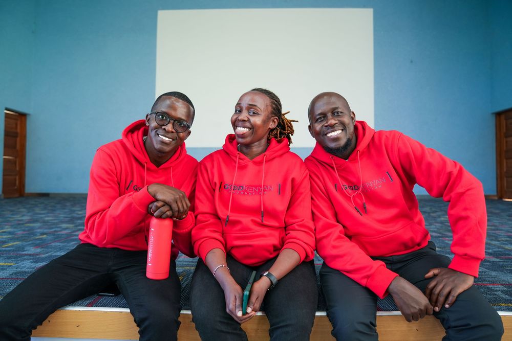 Three men in matching red hoodies sitting together laughing indoors against a blue wall.