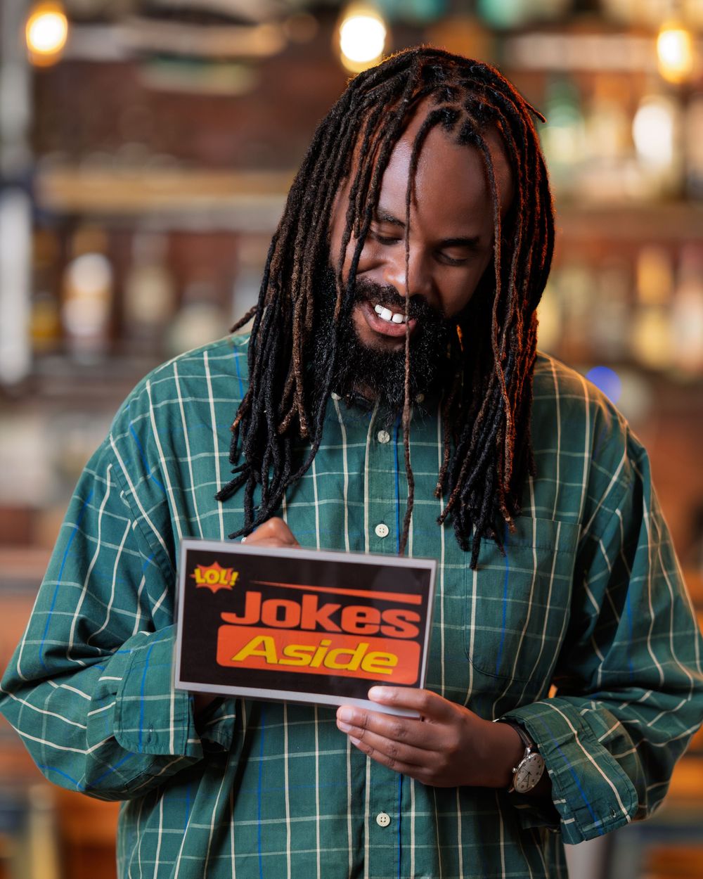 Man with dreadlocks smiling while holding a Jokes Aside comedy show sign indoors.