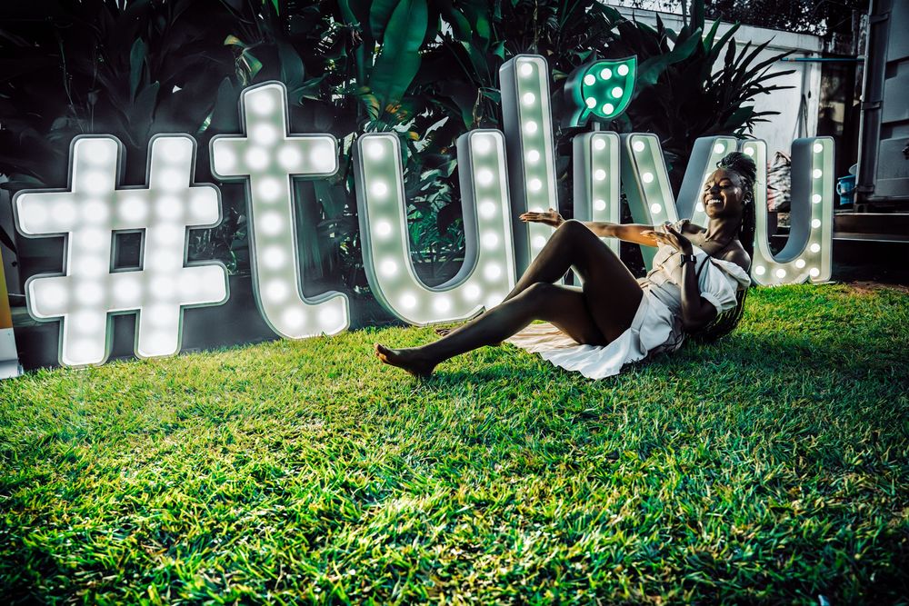 Person posing in front of illuminated #tulivu sign on green grass at night event.