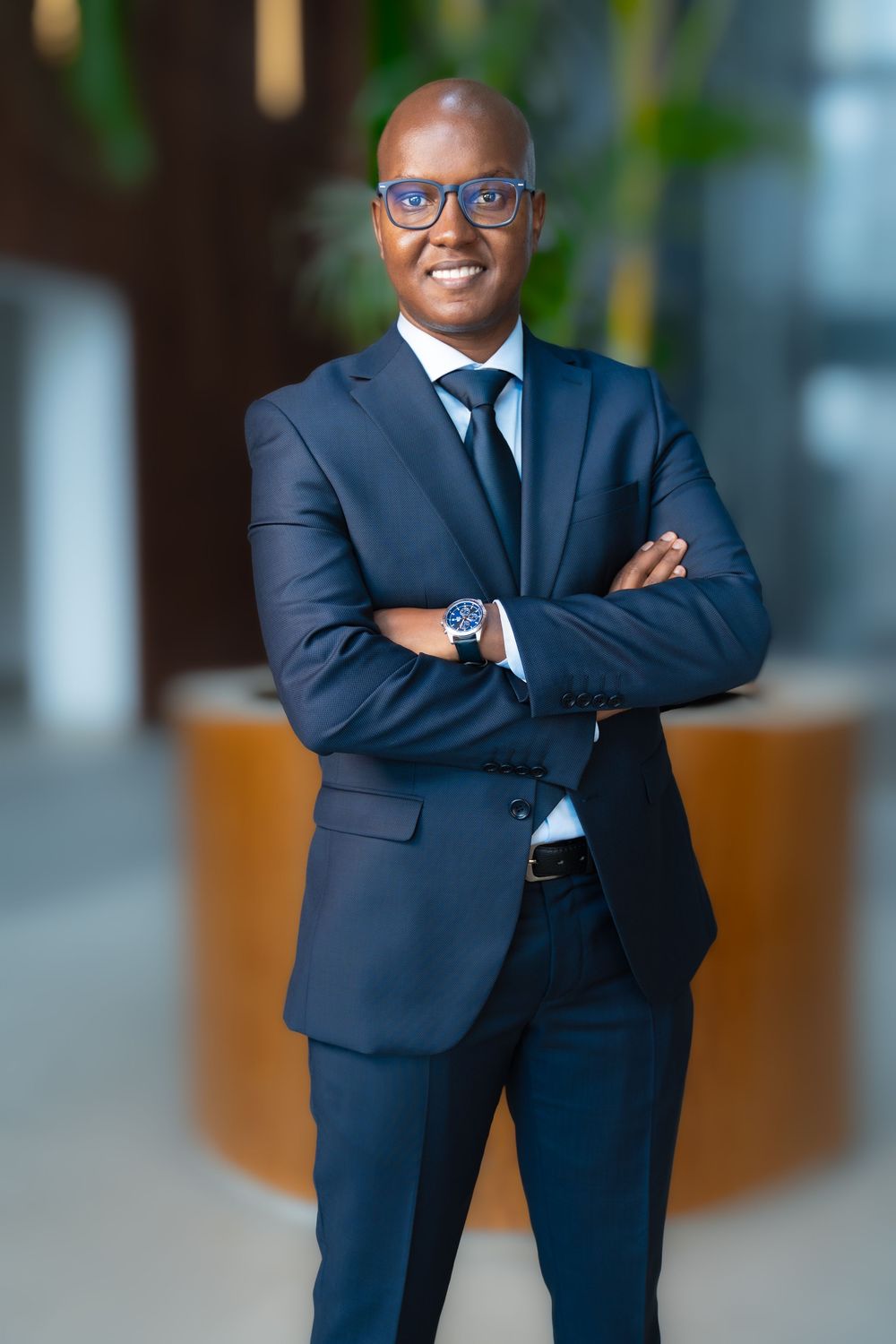 Confident bald African businessman in navy suit with glasses stands arms crossed in modern office.