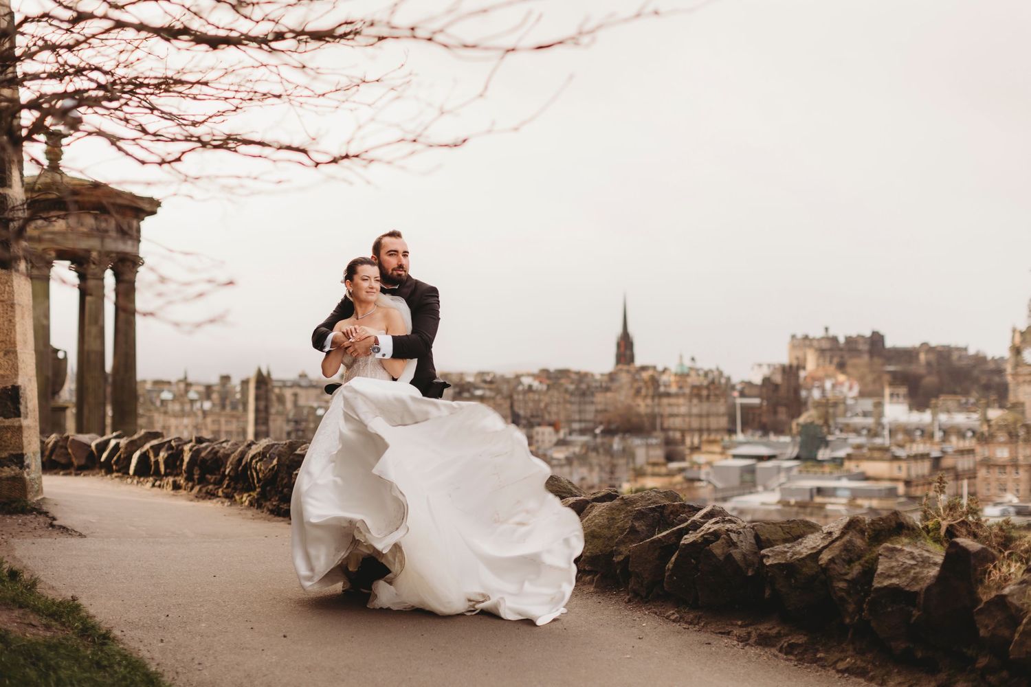 A wedding couple stands on Calton Hill overlooking the Edinburgh cityscape at sunset.