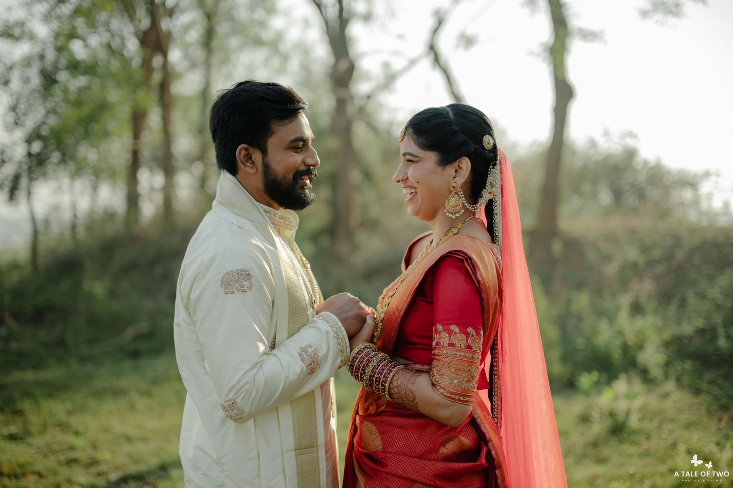 Indian bride in red saree and groom in cream sherwani share a joyful moment outdoors on their wedding day.