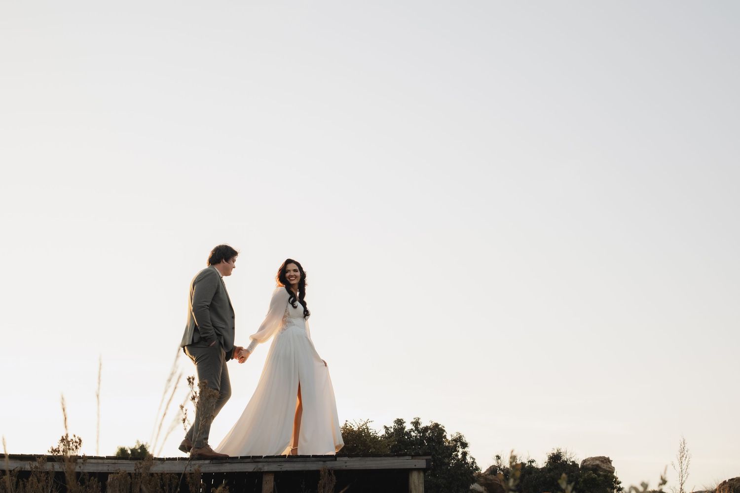 A couple in wedding attire walks through a grassy field at dusk, with the groom carrying a vibrant bouquet of flowers.